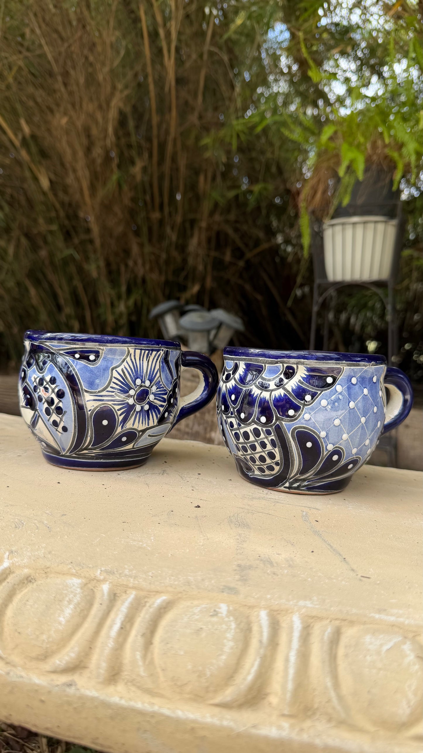 Two blue and white hand-painted ceramic teacups with floral and geometric patterns, resting on a light-colored stone ledge with a bamboo garden background.