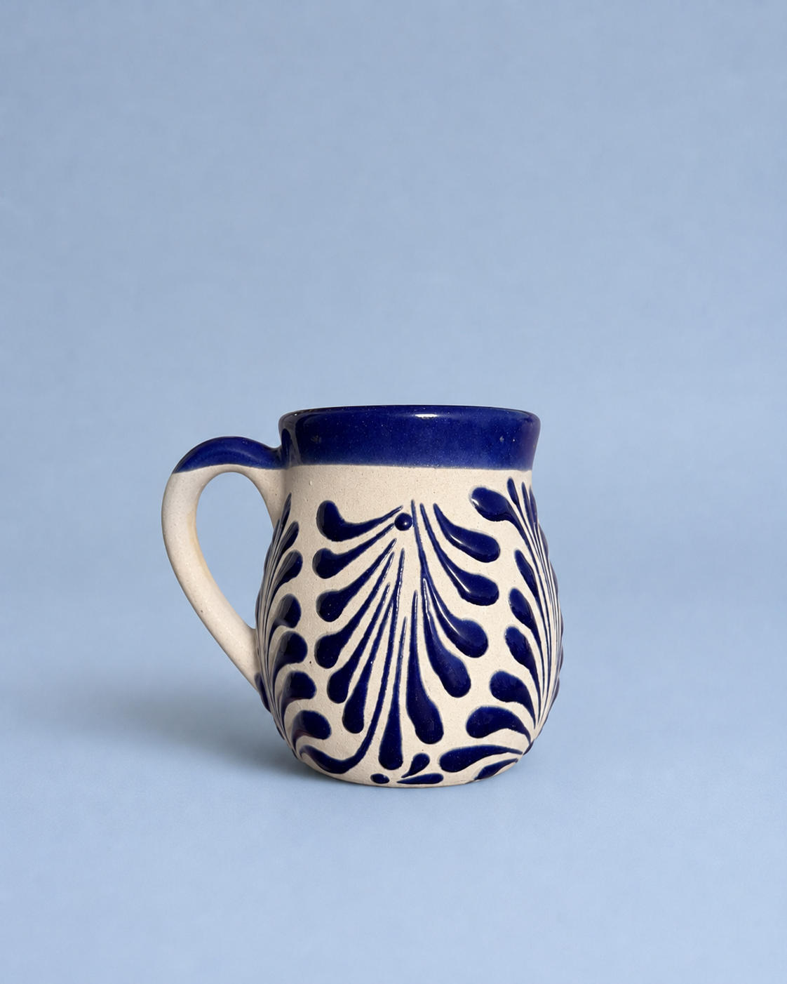 A small ceramic mug with a cream-colored body, a dark blue handle and rim, and a navy blue leaf-like pattern on the front, set against a solid light blue background.