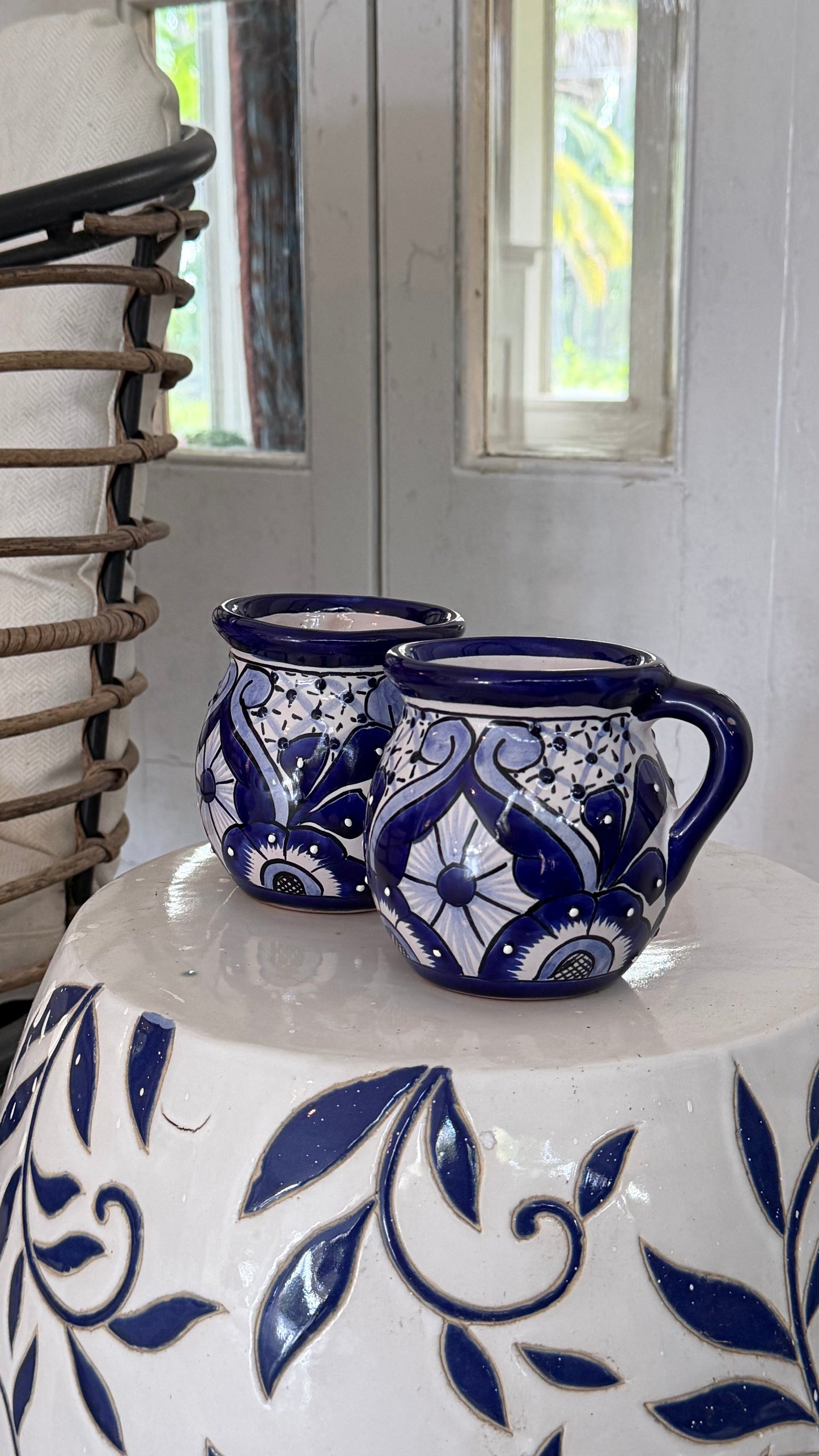 Two handmade ceramic mugs with blue and white floral patterns, displayed on a matching white and blue patterned ceramic pedestal in front of a rustic window.
