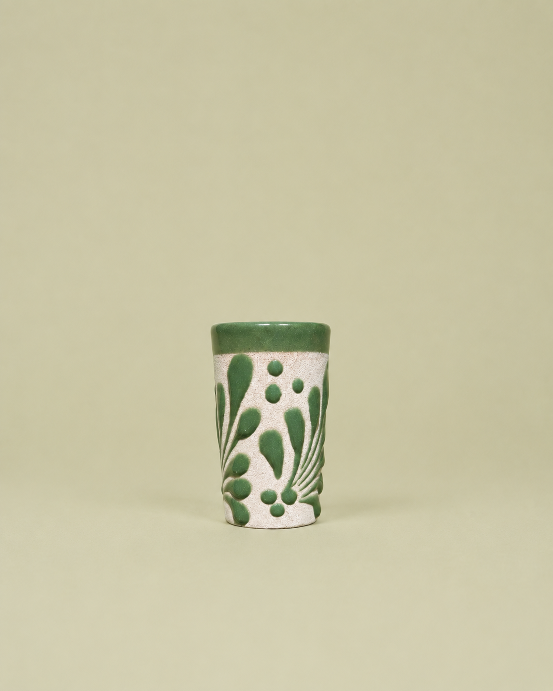 A small cylindrical ceramic shot glass with a textured white body, decorated with green leaf and dot patterns, and finished with a solid green rim, set against a solid light beige background.