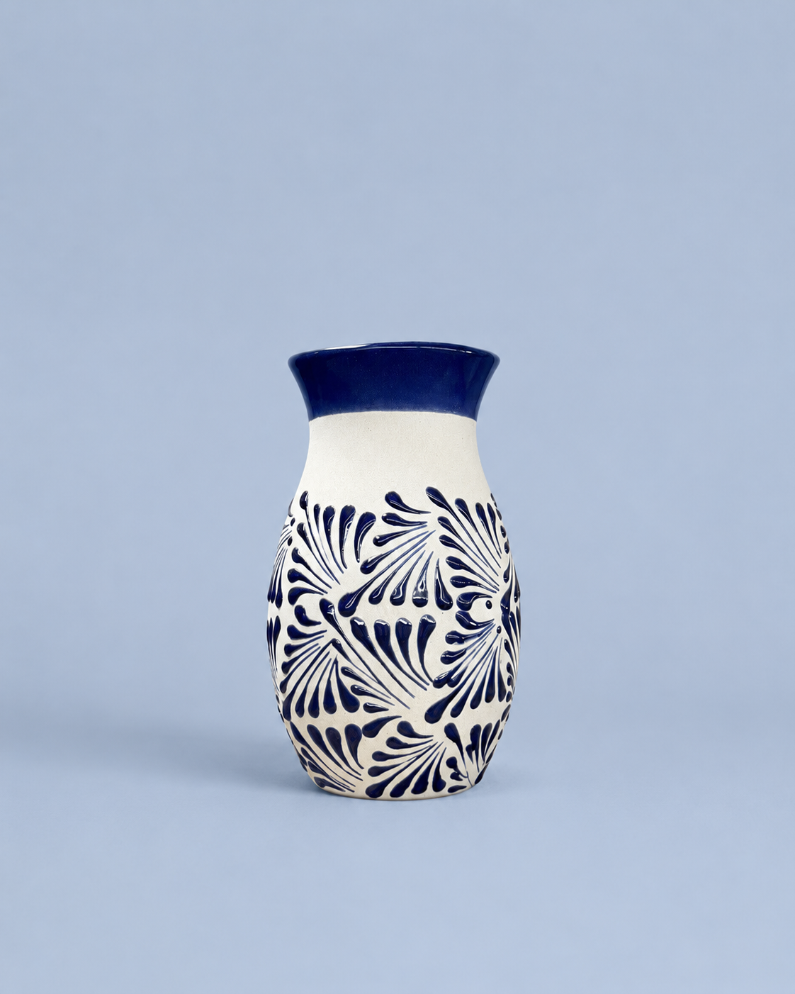 A small ceramic vase with a cream body, dark blue patterned floral design, and a solid dark blue rim, set against a solid light blue background.