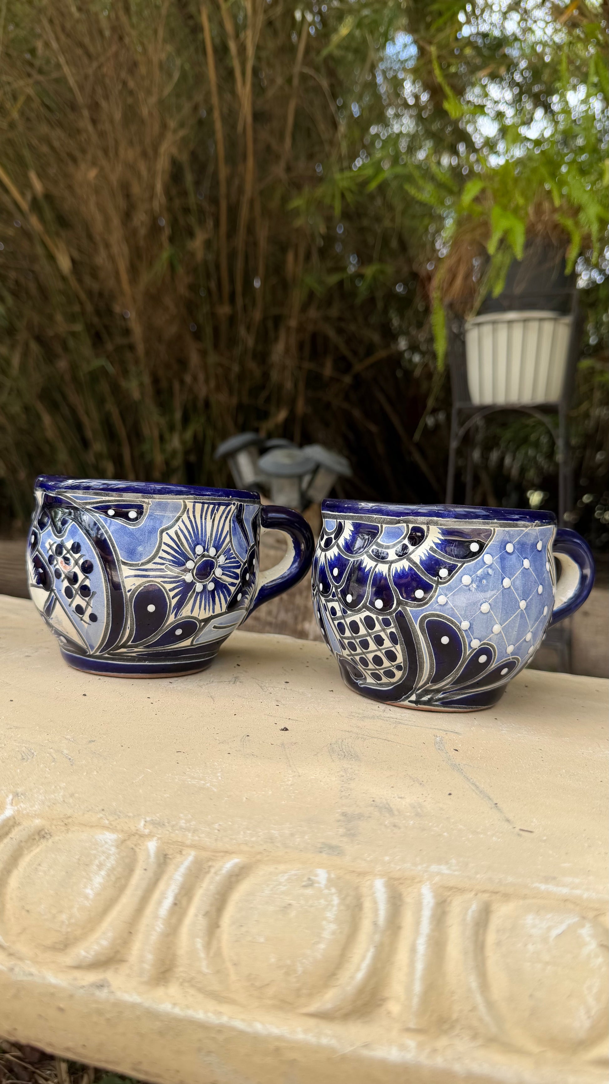 Two blue and white hand-painted ceramic teacups with floral and geometric patterns, resting on a light-colored stone ledge with a bamboo garden background.