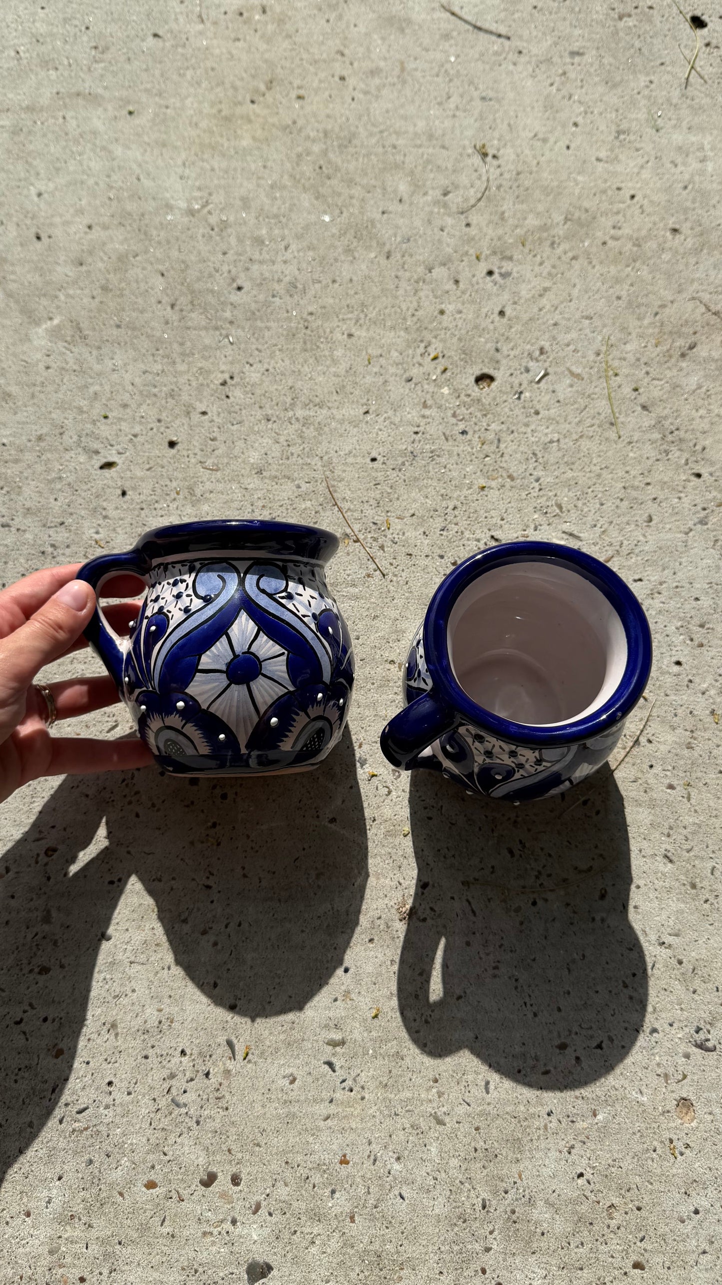 Two handmade ceramic mugs with blue and white floral patterns, one being held by a hand and the other showing its interior, placed on a concrete surface.