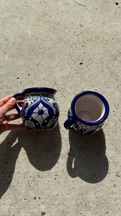 Two handmade ceramic mugs with blue and white floral patterns, one being held by a hand and the other showing its interior, placed on a concrete surface.