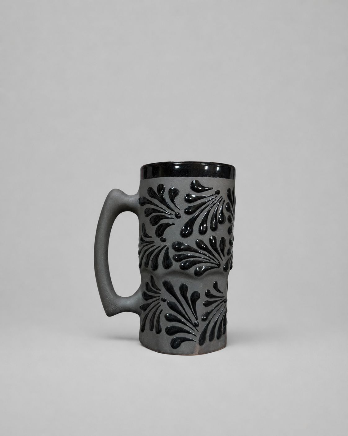 A dark gray ceramic mug with a black handle, black rim, and raised black leaf-like patterns on the body, photographed against a neutral light gray background.
