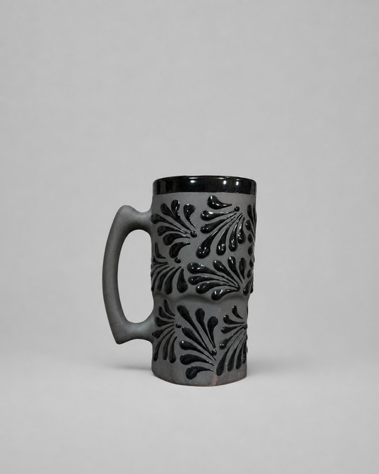 A dark gray ceramic mug with a black handle, black rim, and raised black leaf-like patterns on the body, photographed against a neutral light gray background.