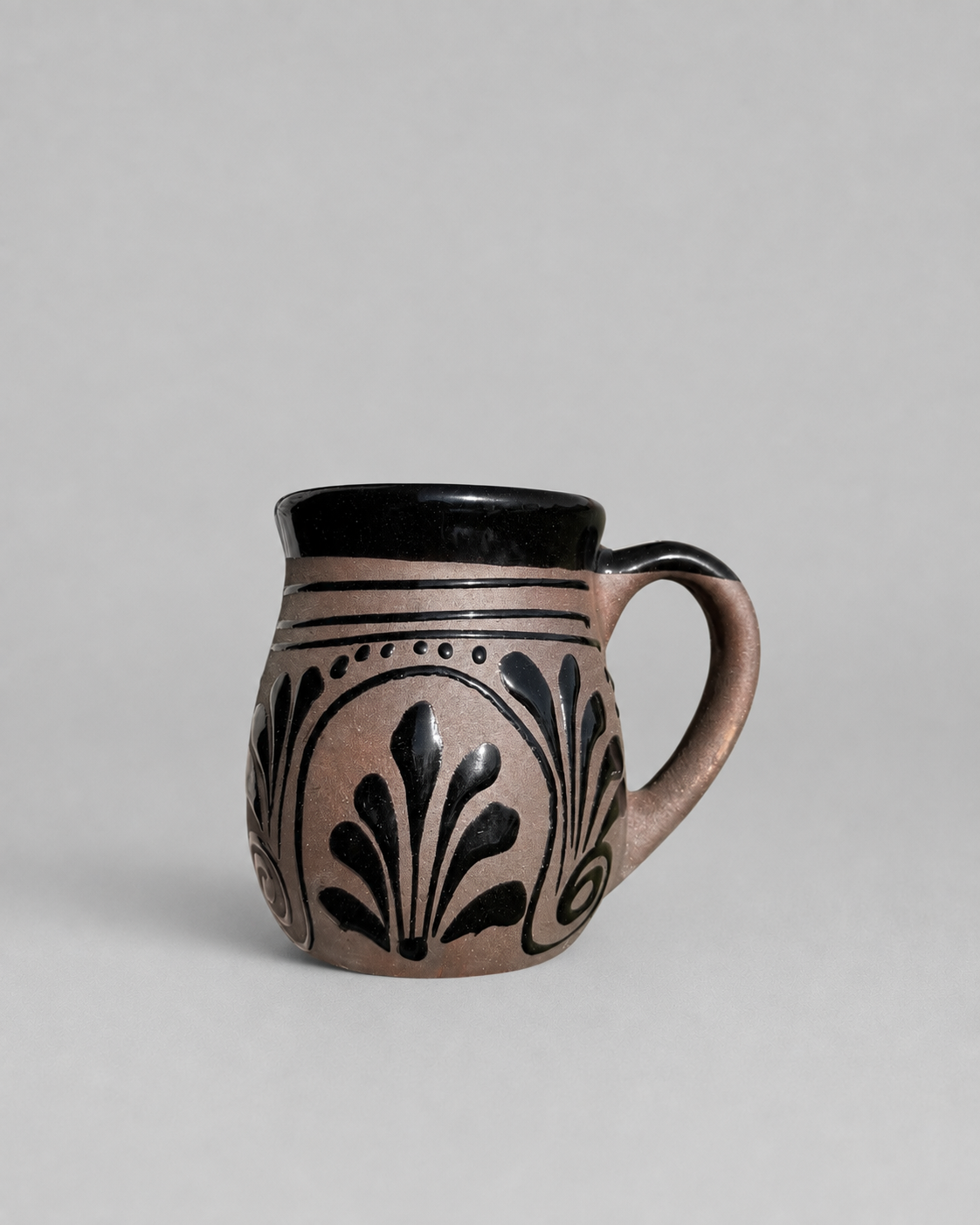 A small ceramic mug with a matte black interior and handle, decorated on the exterior with a dusty pink glaze and black leaf-like patterns, set against a plain light gray background.