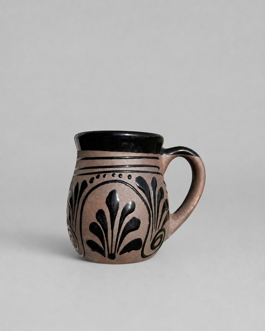 A small ceramic mug with a matte black interior and handle, decorated on the exterior with a dusty pink glaze and black leaf-like patterns, set against a plain light gray background.