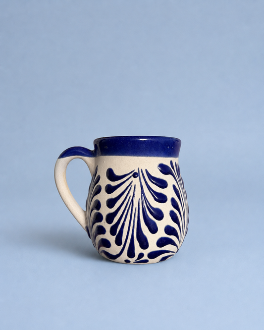 A small ceramic mug with a cream-colored body, a dark blue handle and rim, and a navy blue leaf-like pattern on the front, set against a solid light blue background.