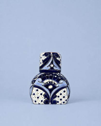 A small decorative ceramic bottle with a matching lid, decorated with a blue and white geometric pattern, set against a solid light blue background.