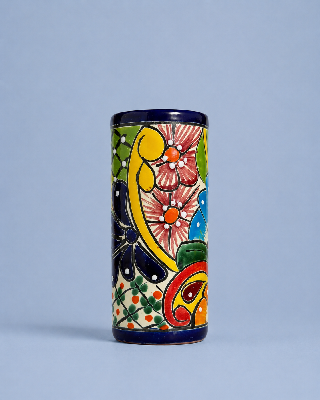 A cylindrical ceramic cup or tumbler with a hand-painted traditional Mexican Talavera style design, featuring a cream background with colorful floral and paisley motifs in yellow, green, pink, blue, and red, finished with a dark blue glazed rim.