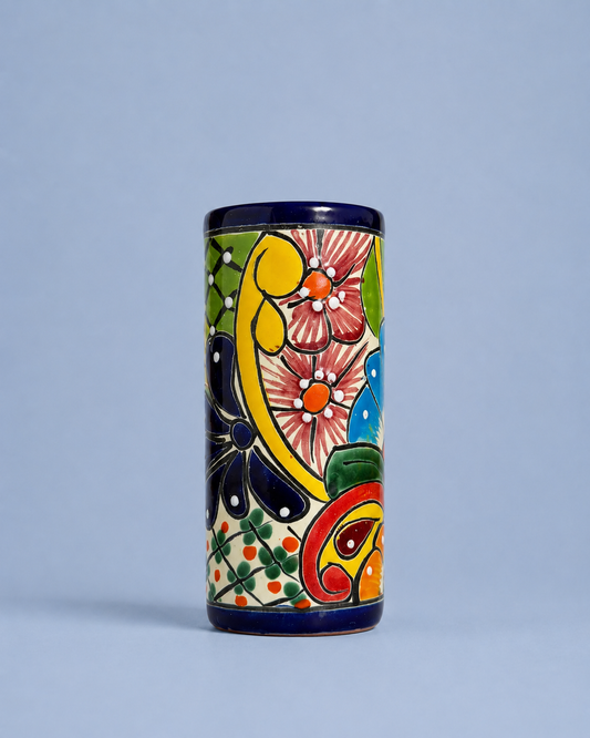 A cylindrical ceramic cup or tumbler with a hand-painted traditional Mexican Talavera style design, featuring a cream background with colorful floral and paisley motifs in yellow, green, pink, blue, and red, finished with a dark blue glazed rim.
