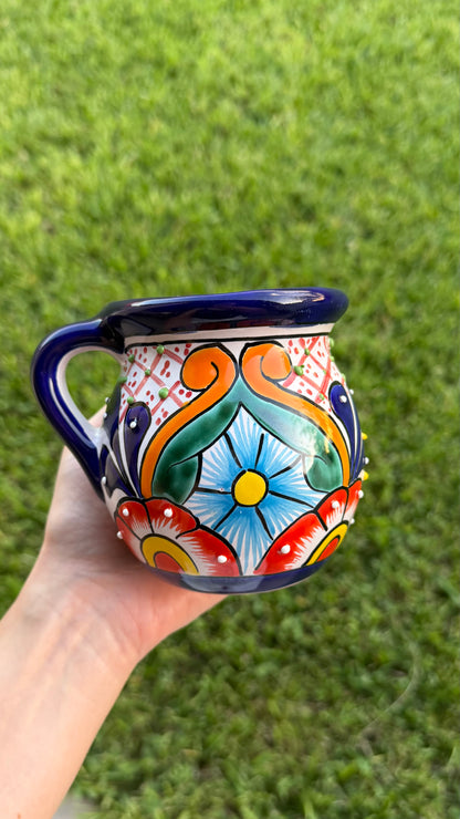 A hand holding a colorful hand-painted ceramic mug outdoors with green grass in the background. The mug features a vibrant Mexican-style floral and scroll pattern in blue, orange, red, green, and white, with a dark blue rim and handle.