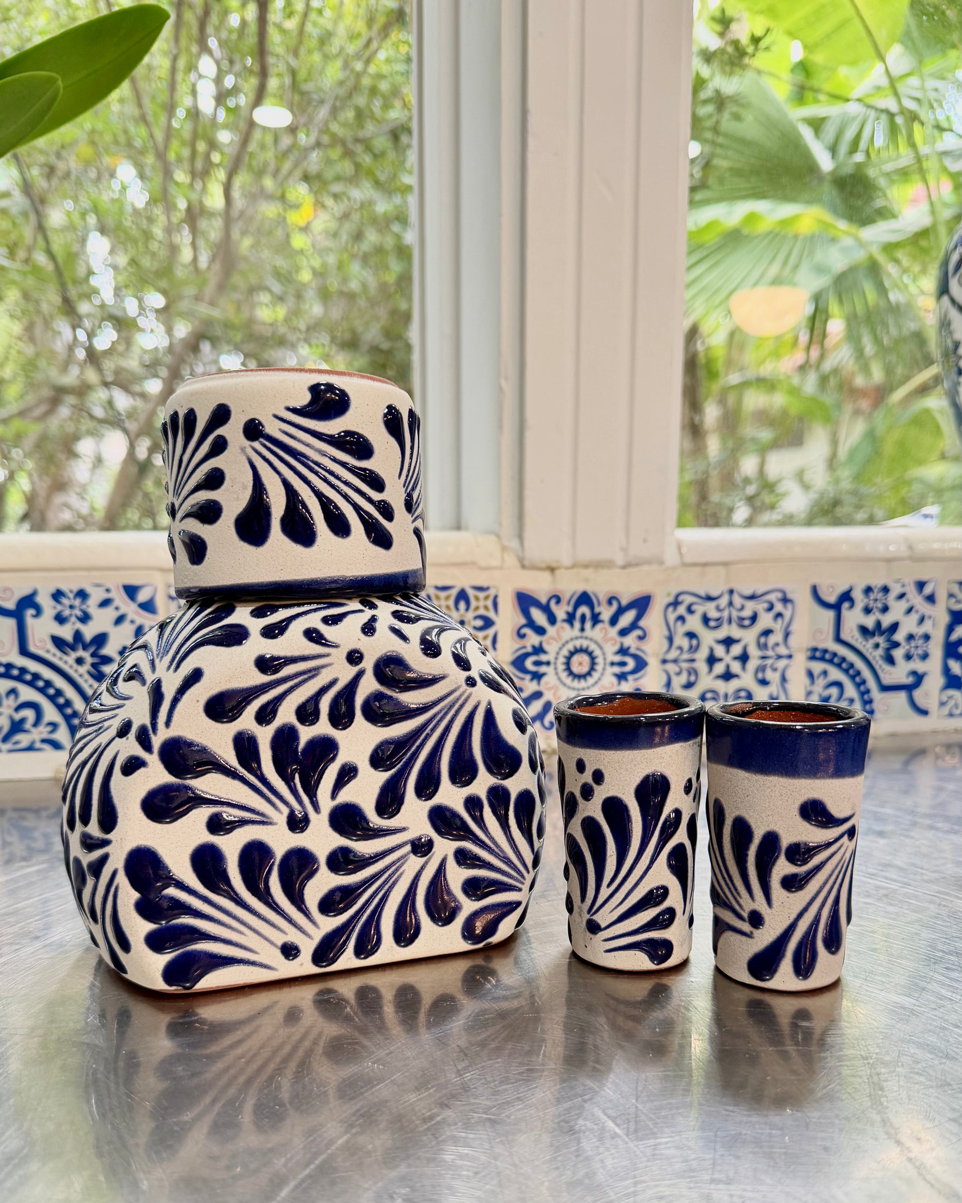 A ceramic set with a large lidded jar and two matching tumblers, decorated with a dark blue hand-painted floral pattern on a white base, set on a reflective metal surface in front of a window.