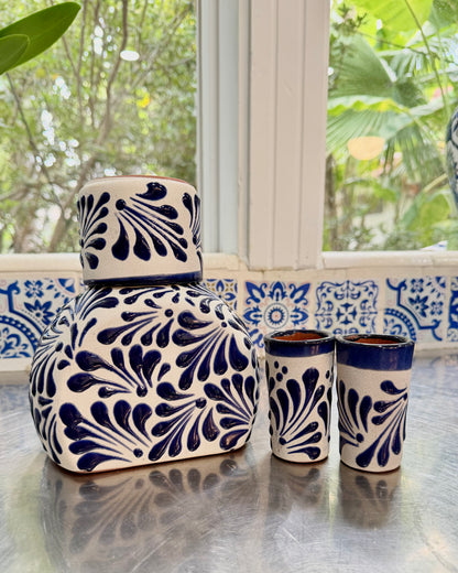 A ceramic set with a large lidded jar and two matching tumblers, decorated with a dark blue hand-painted floral pattern on a white base, set on a reflective metal surface in front of a window.