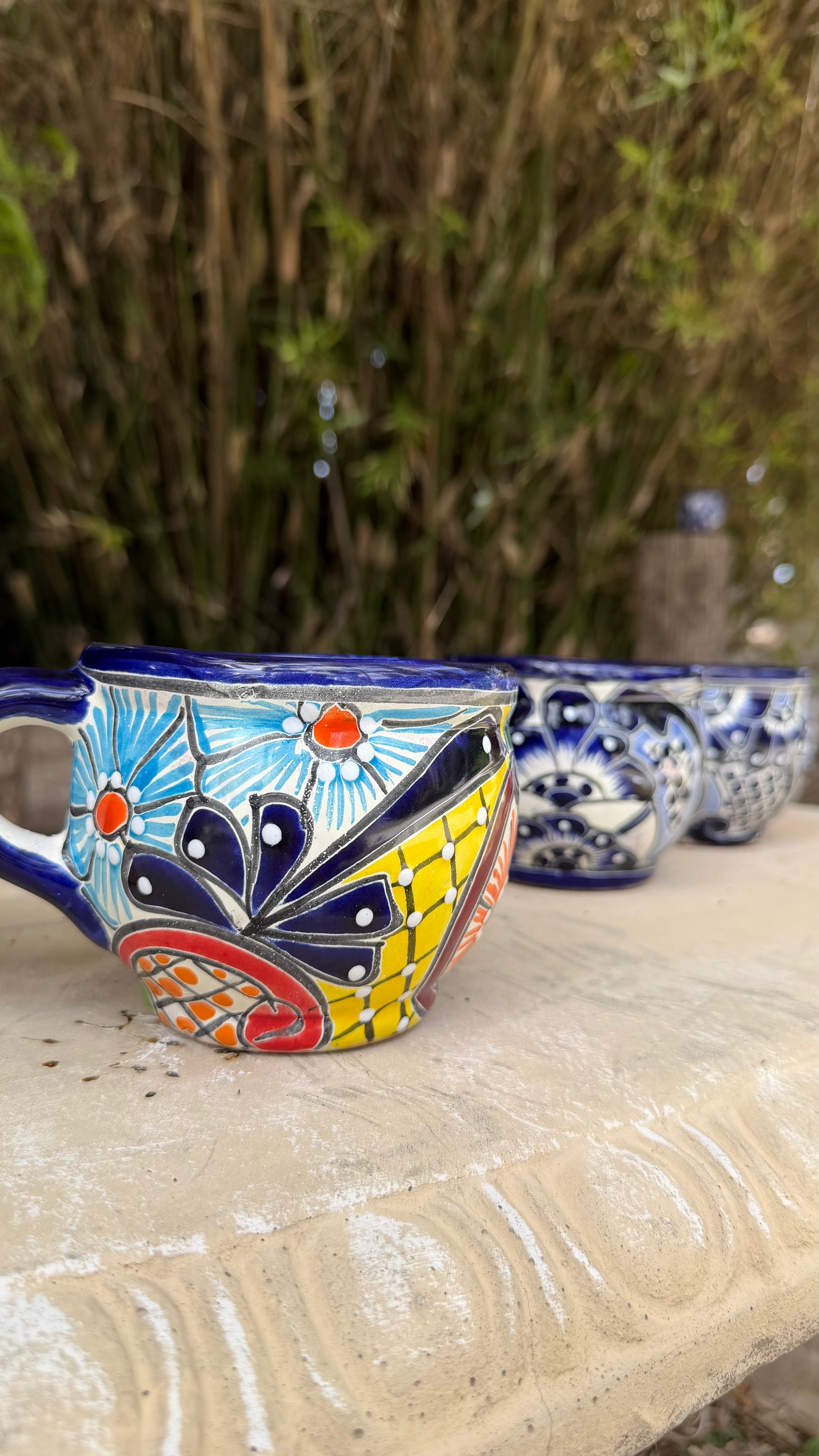 A colorful hand-painted ceramic mug with a blue handle and rim, featuring a vibrant tropical floral and butterfly pattern, sitting on a stone ledge with bamboo in the background.
