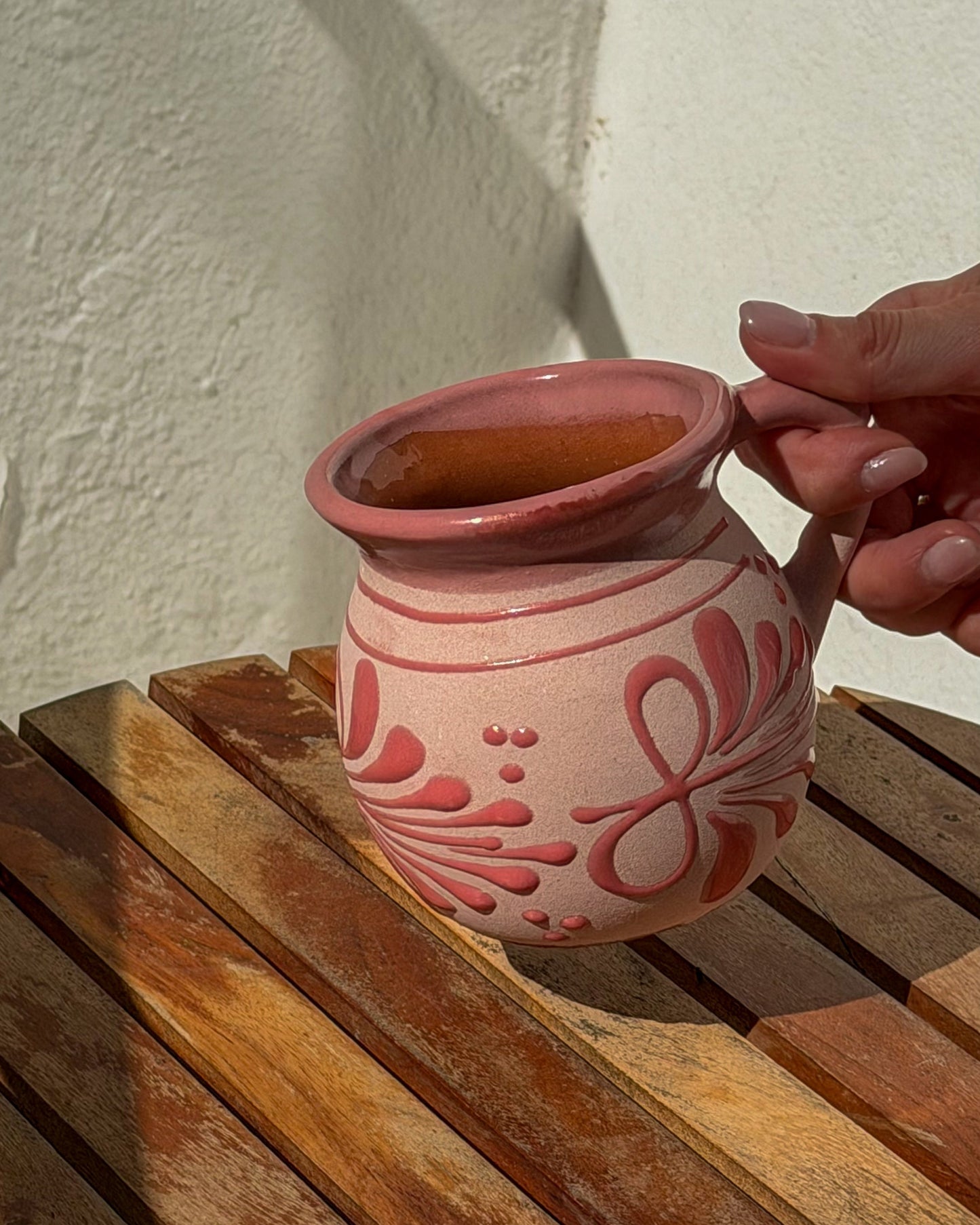 A hand holds a small pink ceramic mug with a floral pattern, sitting on a wooden slatted surface against a textured white wall.