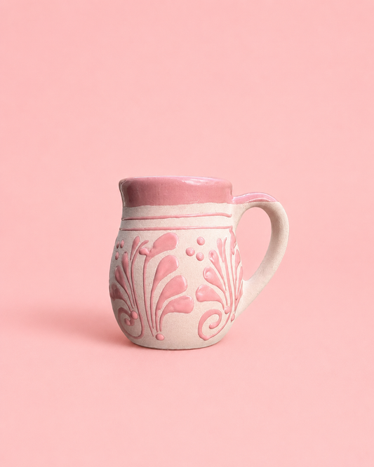 A small pink and off-white ceramic pitcher with embossed floral and scroll patterns, set against a solid pink background.