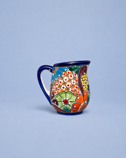 A colorful hand-painted ceramic mug with a blue handle and rim, featuring a traditional Spanish or Mexican floral and geometric pattern, set against a solid light blue background.