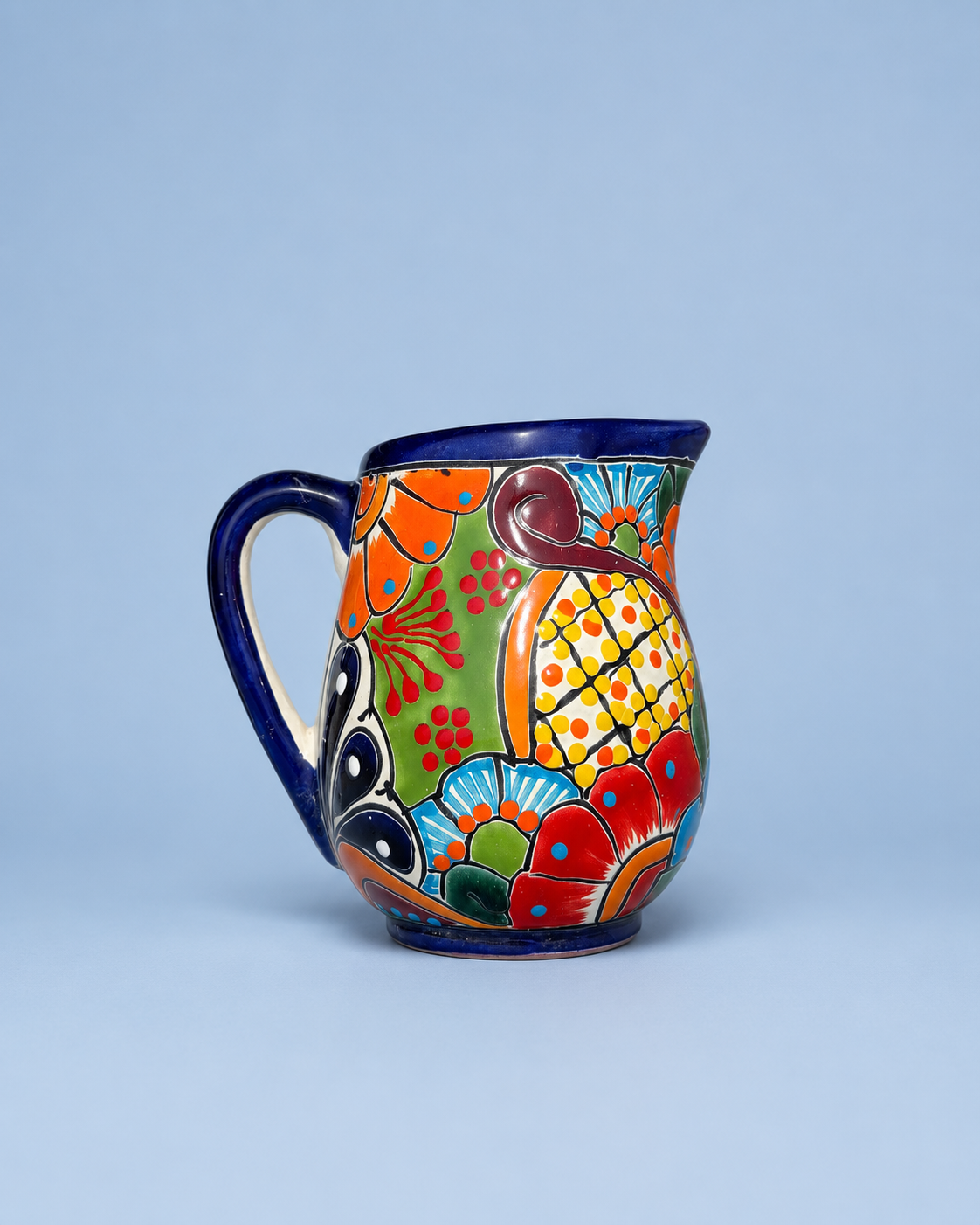 A colorful ceramic pitcher with a hand-painted Mexican Talavera style design, featuring a blue handle and rim, set against a solid light blue background.