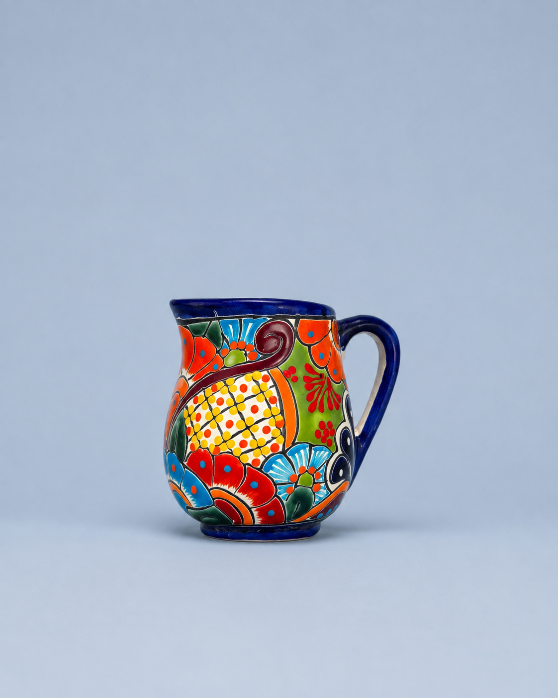 A small ceramic pitcher with a colorful hand-painted Mexican Talavera style design, featuring a blue cobalt rim, handle, and base, set against a solid light blue-gray background.