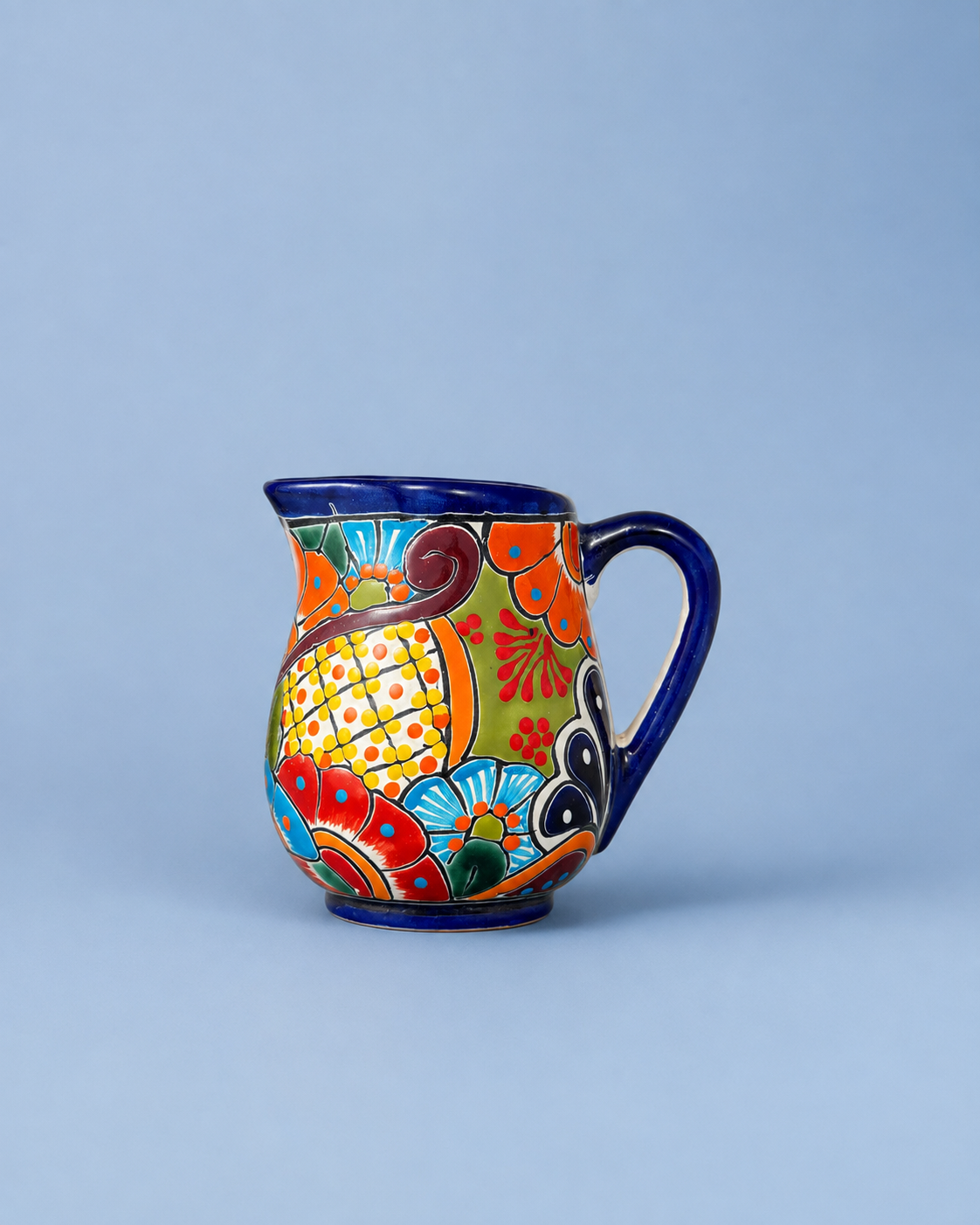 A colorful hand-painted ceramic pitcher with a dark blue handle and rim, decorated with vibrant orange, yellow, red, and blue floral and geometric patterns, set against a solid light blue background.