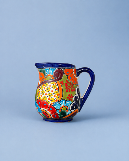 A colorful hand-painted ceramic pitcher with a dark blue handle and rim, decorated with vibrant orange, yellow, red, and blue floral and geometric patterns, set against a solid light blue background.