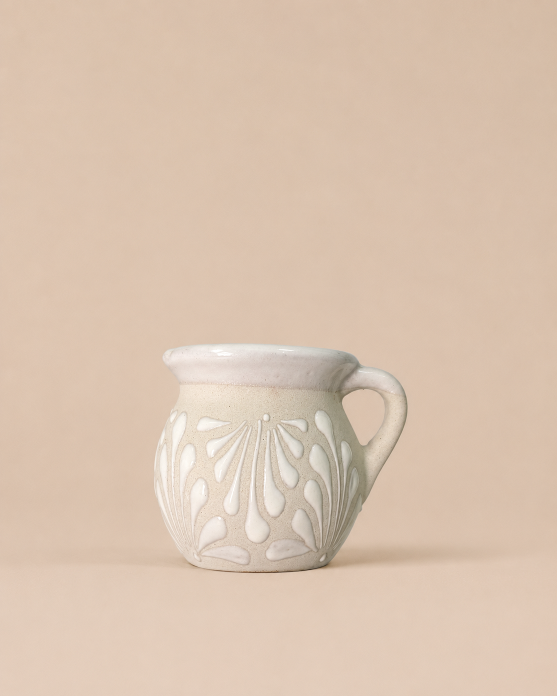 A small ceramic pitcher with a textured, speckled off-white glaze and embossed leaf patterns, set against a solid beige background.