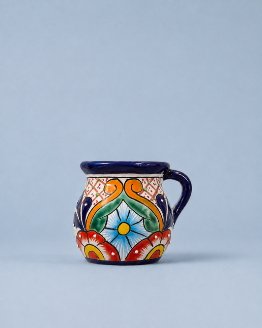 A small hand-painted ceramic mug with a handle, featuring a traditional Mexican Talavera style design with a blue rim, handle, and base, and a colorful floral and scroll pattern on a light gray background.