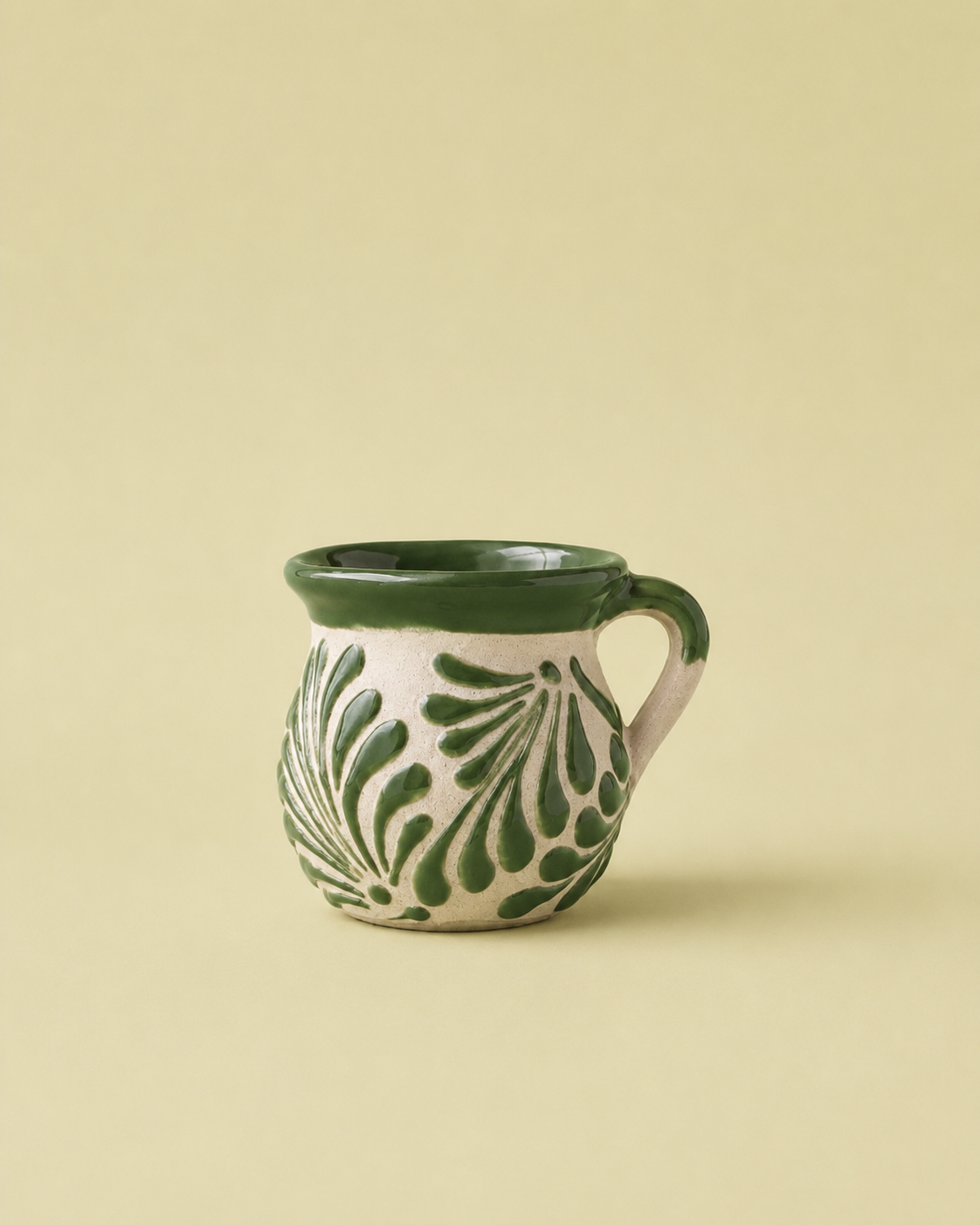 A small ceramic mug with a dark green rim, handle, and leaf pattern on an off-white body, set against a solid pale yellow background.