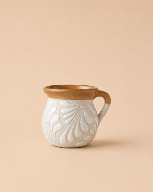 A small ceramic mug with a textured, embossed floral pattern on a warm off-white body, featuring a contrasting glazed brown rim and handle, set against a solid beige background.