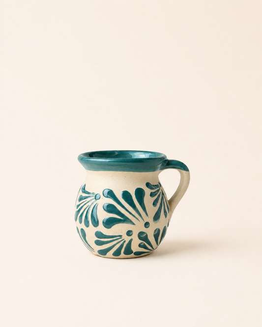 A small ceramic mug with a teal interior, rim, and handle, decorated with a teal floral pattern on an off-white exterior, set against a solid cream background.