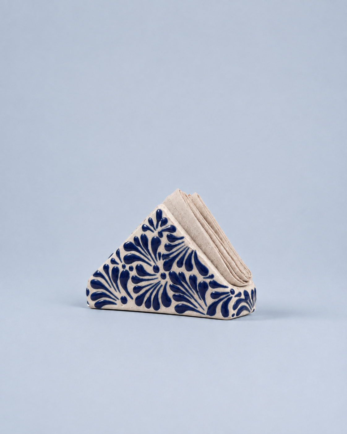 A triangular stack of beige paper napkins with a navy blue floral pattern on the front, set against a solid light blue-gray background.