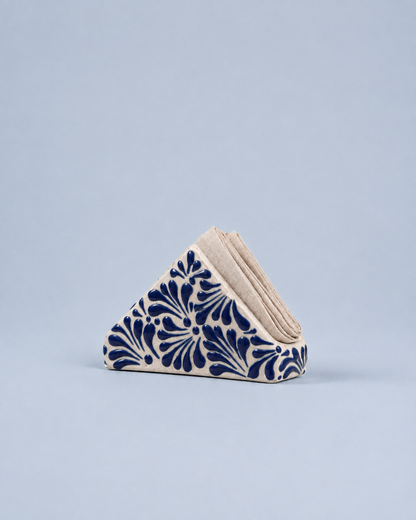 A triangular stack of beige paper napkins with a navy blue floral pattern on the front, set against a solid light blue-gray background.