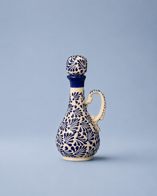 A ceramic oil bottle with a matching stopper, decorated with a blue and cream floral pattern, set against a solid light blue background.