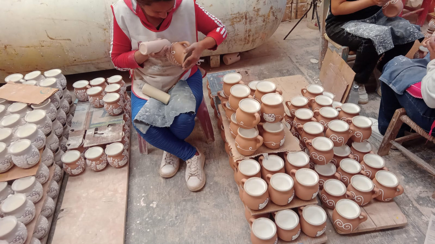 A person in a workshop shapes a ceramic cup while stacks of unglazed clay cups and small bowls are arranged on tables around them.