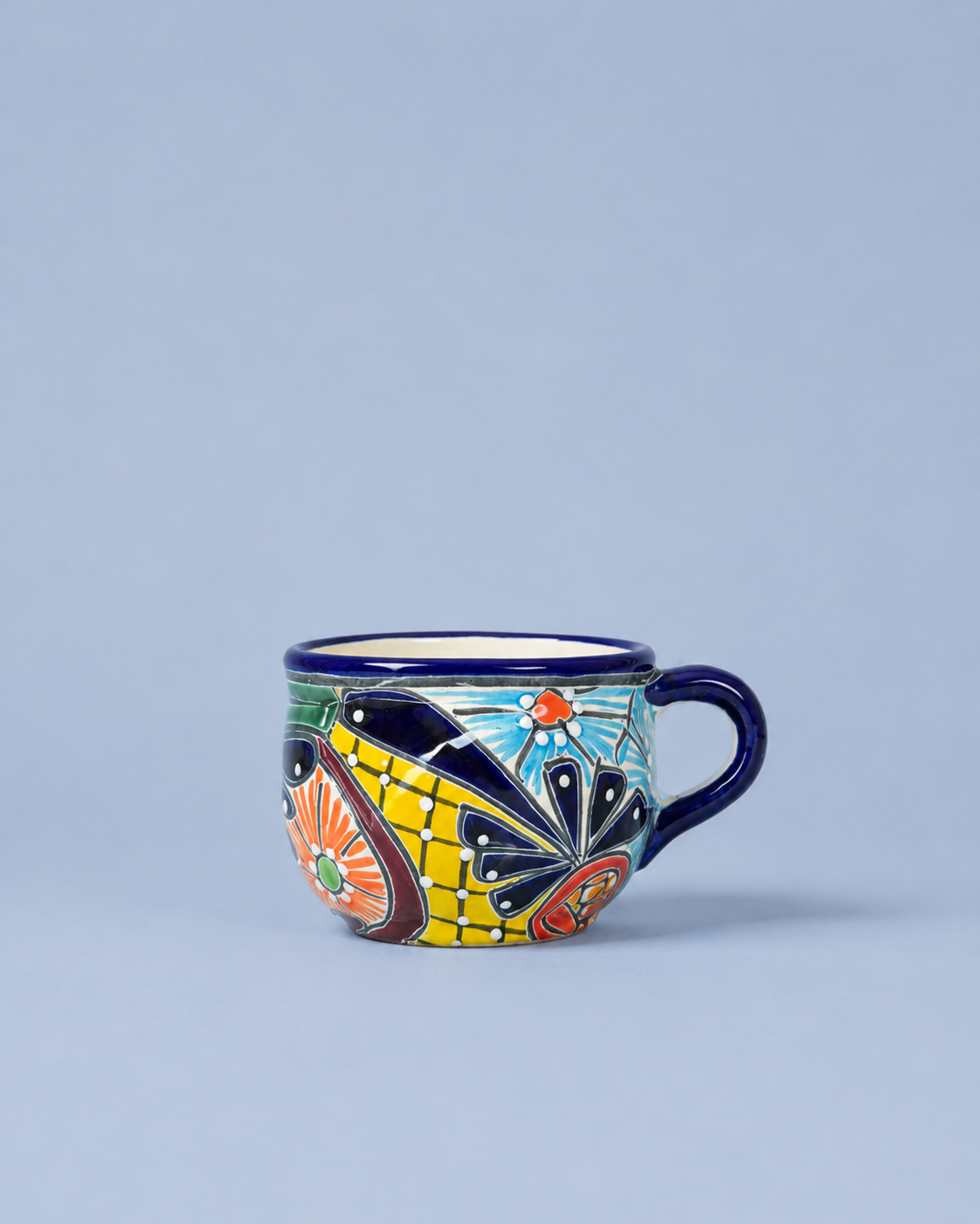 A small ceramic coffee cup with a colorful hand-painted pattern featuring yellow, blue, orange, and black, set against a solid light blue-gray background.