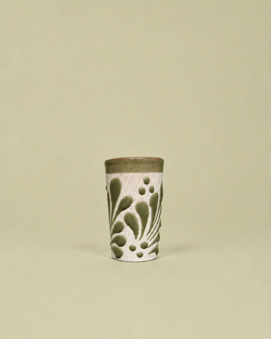 A small cylindrical ceramic shot glass with a textured white body, olive green leaf-like patterns, and a solid olive green rim, set against a solid light beige background.