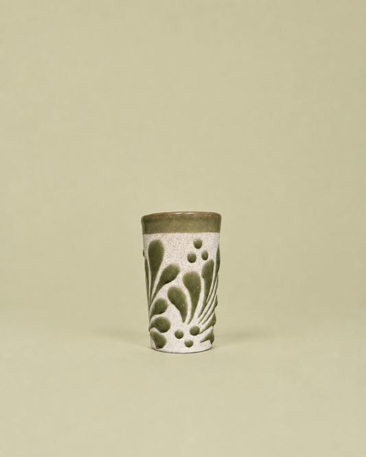 A small cylindrical ceramic shot glass with a textured white body, olive green leaf-like patterns, and a solid olive green rim, set against a solid light beige background.