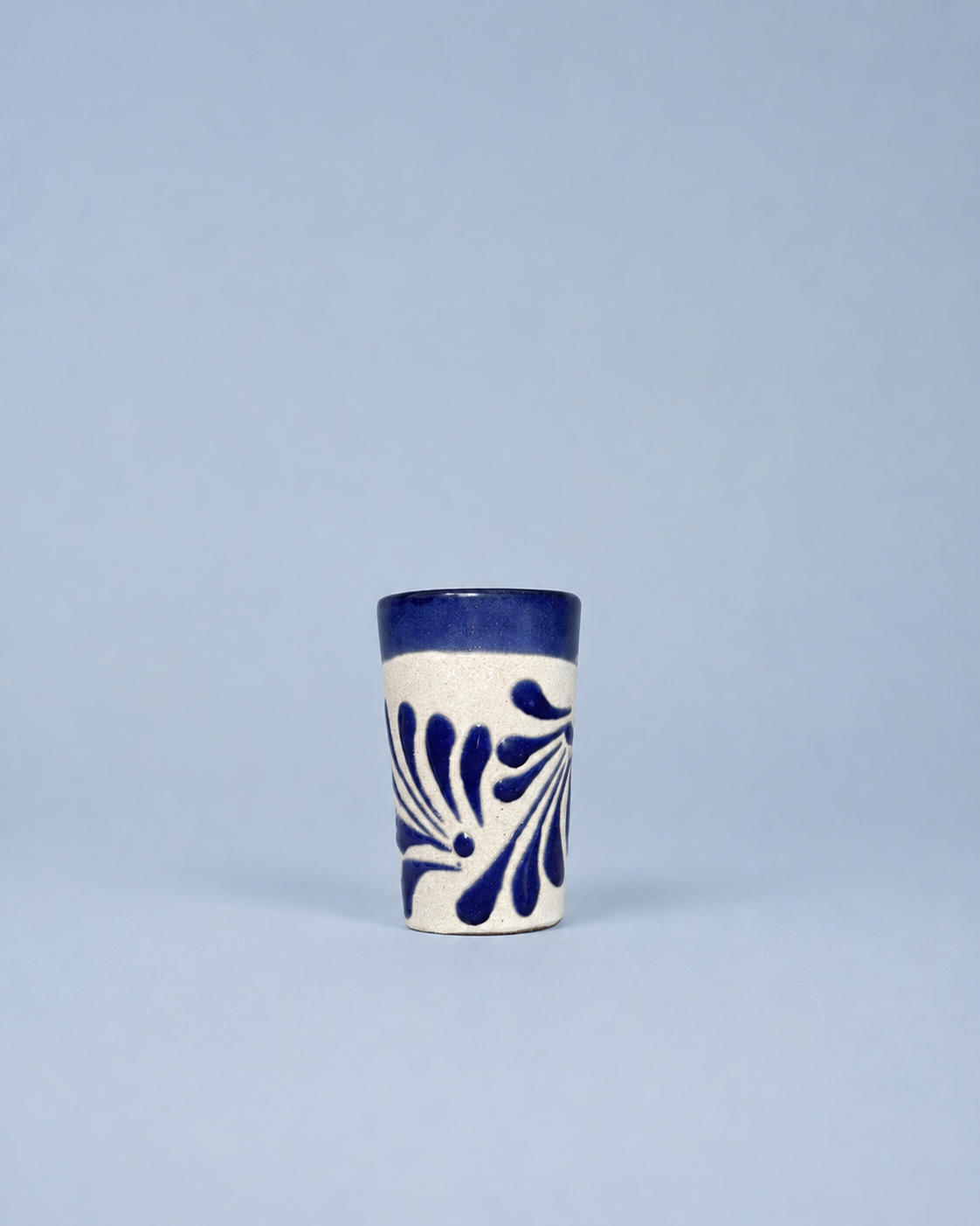 A small ceramic shot glass with a white body, dark blue patterned leaf design, and a solid dark blue rim, set against a solid light blue background.