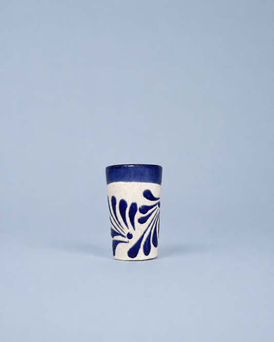 A small ceramic shot glass with a white body, dark blue patterned leaf design, and a solid dark blue rim, set against a solid light blue background.