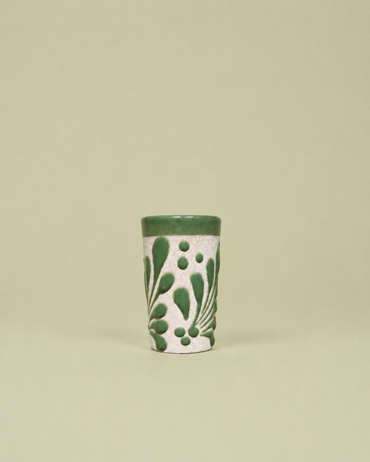 A small cylindrical ceramic shot glass with a textured white body, decorated with green leaf and dot patterns, and finished with a solid green rim, set against a solid light beige background.