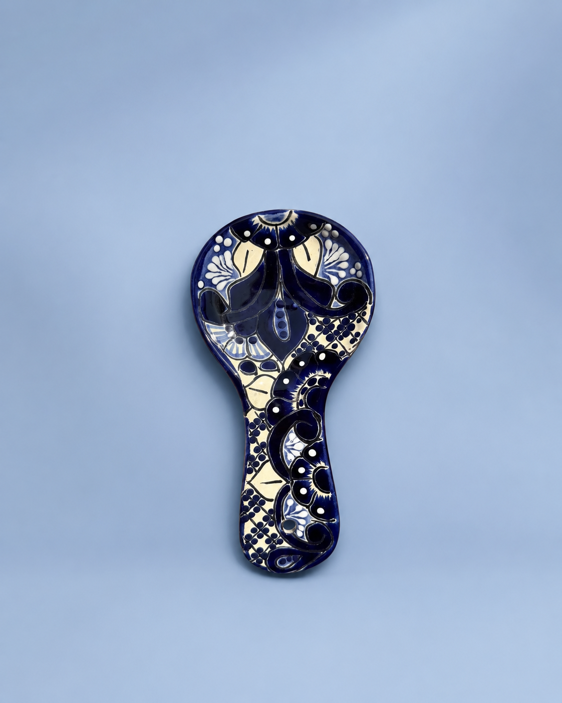 A decorative ceramic spoon or small dish with a handle, featuring an intricate hand-painted floral and geometric pattern in deep navy blue, cream, and white, set against a soft light blue background.