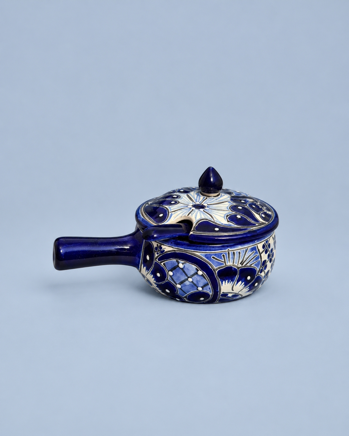 A small cobalt blue ceramic lidded pot or teapot with ornate white and gold floral patterns, set against a solid light blue-gray background.