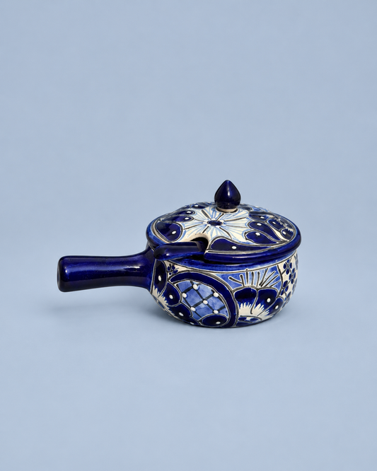 A small cobalt blue ceramic lidded pot or teapot with ornate white and gold floral patterns, set against a solid light blue-gray background.