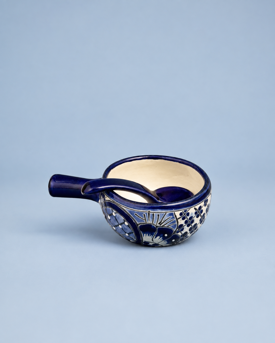 A small ceramic cup with a handle, featuring a deep cobalt blue exterior with white floral and geometric patterns, and a cream-colored interior, set against a solid light blue background.