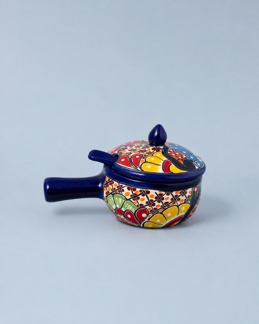 A small hand-painted ceramic lidded pot with a dark blue handle and lid, decorated with a colorful floral and geometric pattern in red, yellow, green, and white, set against a solid light gray background.