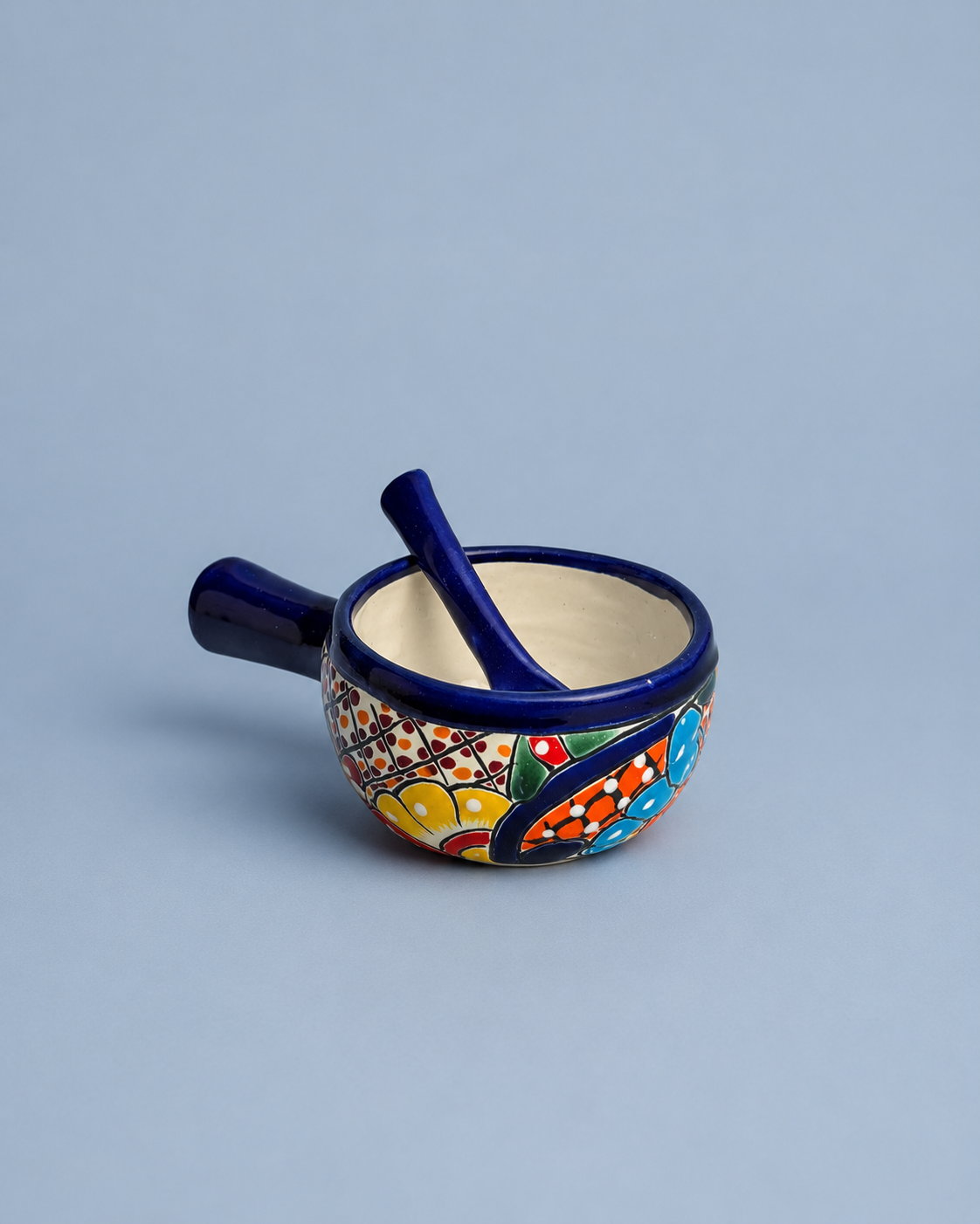 A small ceramic mortar and pestle set with a dark blue handle and rim, decorated with a colorful hand-painted floral pattern on the side, set against a solid light blue-gray background.
