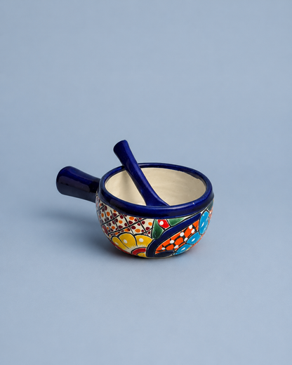 A small ceramic mortar and pestle set with a dark blue handle and rim, decorated with a colorful hand-painted floral pattern on the side, set against a solid light blue-gray background.