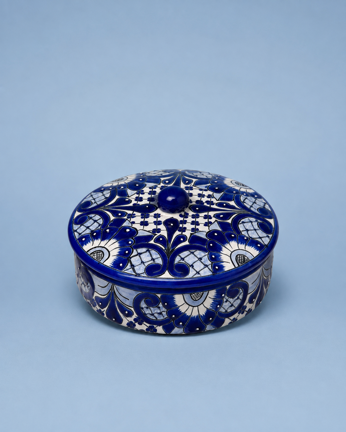 A round lidded ceramic box with intricate cobalt blue and white floral patterns, photographed on a solid light blue background.